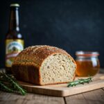 loaf of bread on cutting board with sprigs of rosemary in front and bottle of bear in left background and honey jar in the right background