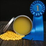 open tin of yellow furniture polish on table with lid to the left, blue ribbon to the right and pile of beeswax pellets on the table to the left of tin