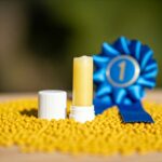 yellow lip balm with lid to the left, blue 1st place ribbon to right surrounded by yellow beeswax pellets