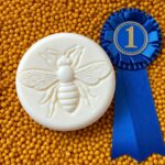 Round white lotion bar with embossed bee on the front resting on yellow beeswax pellets with 1st place blue ribbon to the right