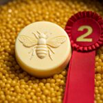 round yellow lotion bar with bee embossed on the front on a field of yellow beeswax with 2nd place red ribbon to the right of lotion bar