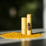 table of beeswax pellets with yellow tube of lip balm standing upright with lid off and honey bee on the front of the tube